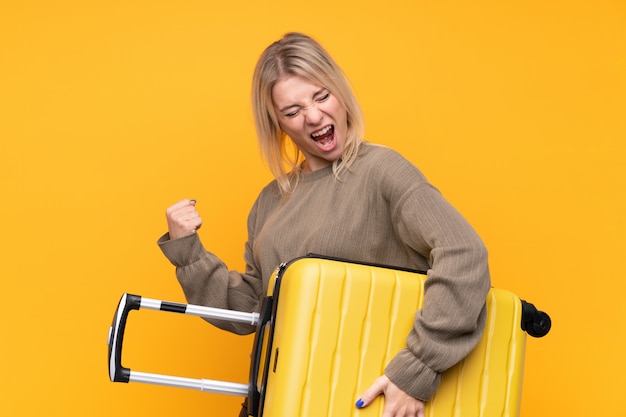 Молодая белокурая женщина над изолированной желтой стеной в каникулах с чемоданом перемещения
