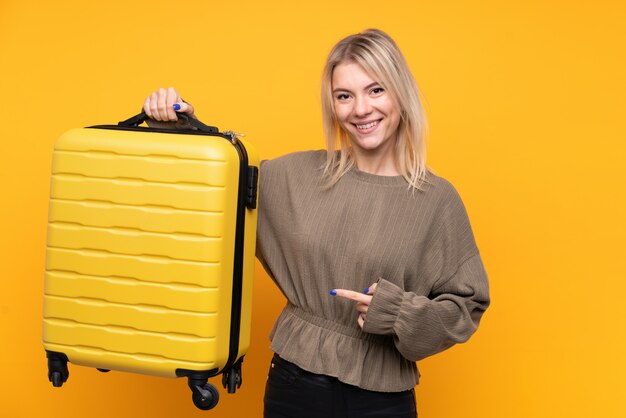 Young blonde woman over isolated yellow wall in vacation with travel suitcase