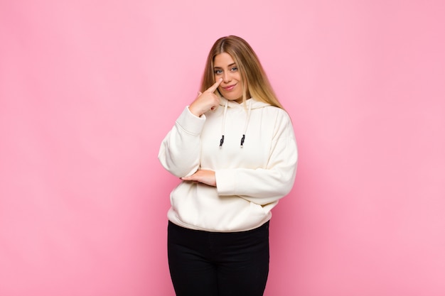 Young blonde woman keeping an eye on you, not trusting, watching and staying alert and vigilant against wall