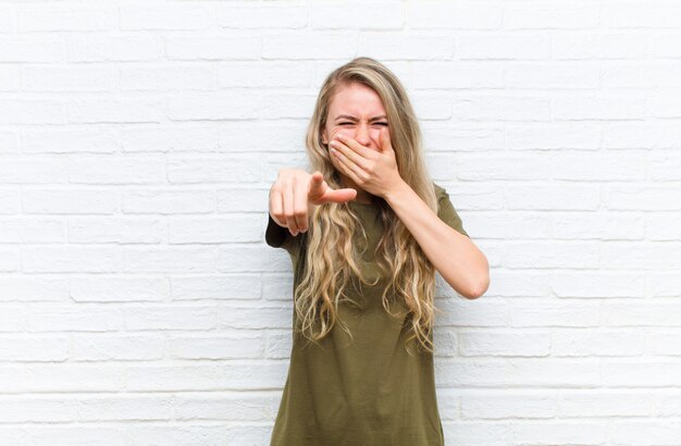 Young blonde woman laughing at you, pointing to and making fun of or mocking you against brick wall