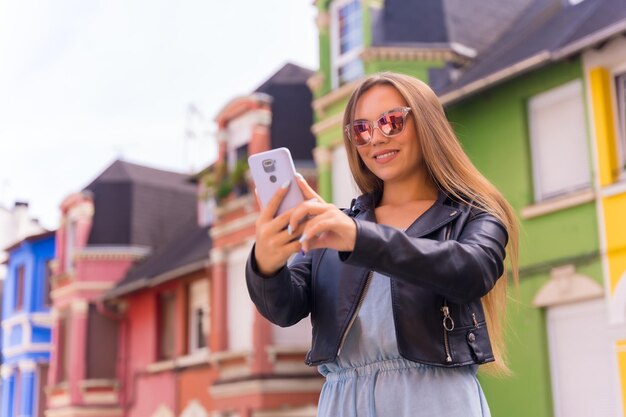 Молодая блондинка в кожаной куртке и солнцезащитных очках улыбается с мобильным телефоном за красочным фасадом
