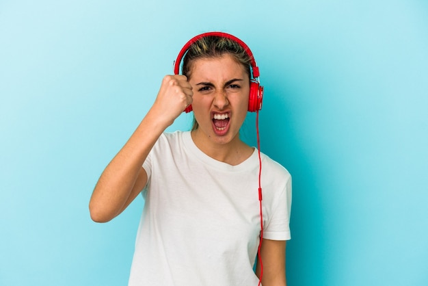 Young blonde woman listening to music on headphones isolated on blue wall showing fist, aggressive facial expression.