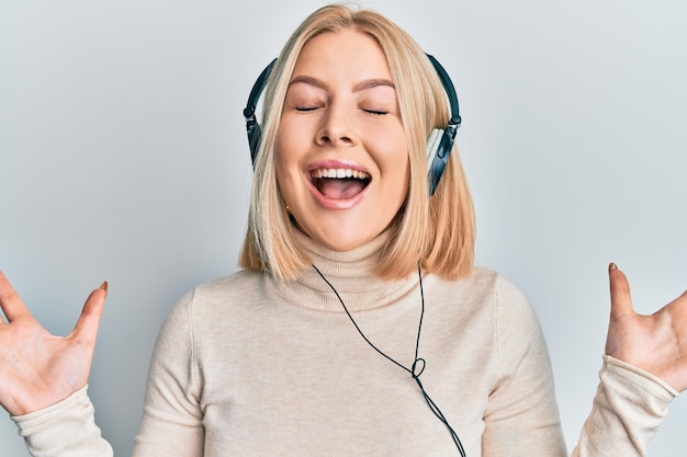 Young blonde woman listening to music using headphones celebrating mad and crazy for success with arms raised and closed eyes screaming excited winner concept