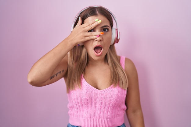 Young blonde woman listening to music using headphones peeking in shock covering face and eyes with hand, looking through fingers with embarrassed expression.