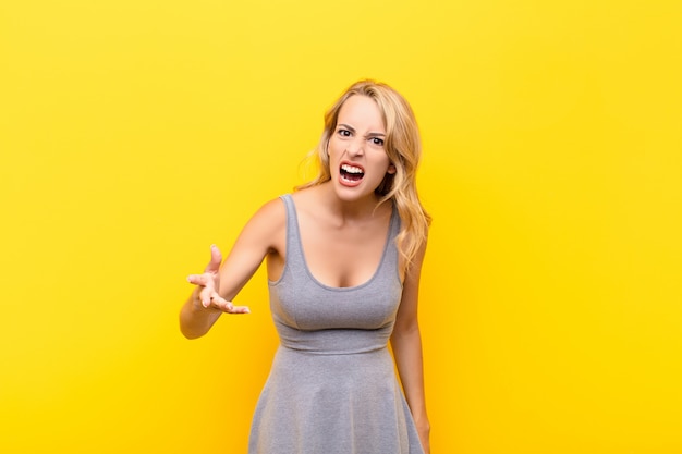 Young blonde woman looking angry, annoyed and frustrated screaming wtf or what's wrong with you against orange wall