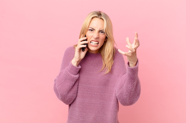 Young blonde woman looking angry, annoyed and frustrated and talking with a smartphone