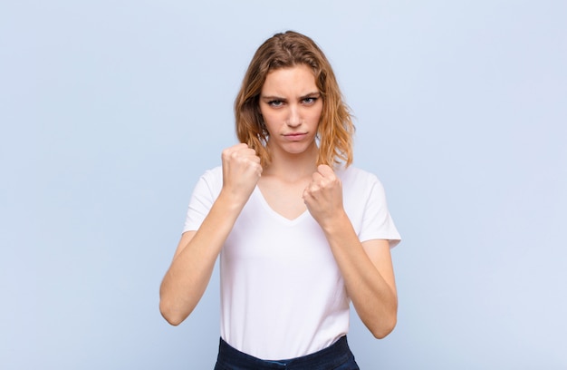 Young blonde woman looking confident, angry, strong and aggressive, with fists ready to fight in boxing position against flat color wall