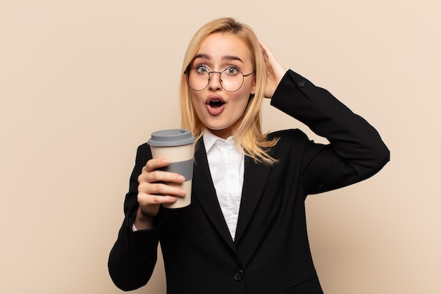 Young blonde woman looking excited and surprised, open-mouthed with both hands on head, feeling like a lucky winner