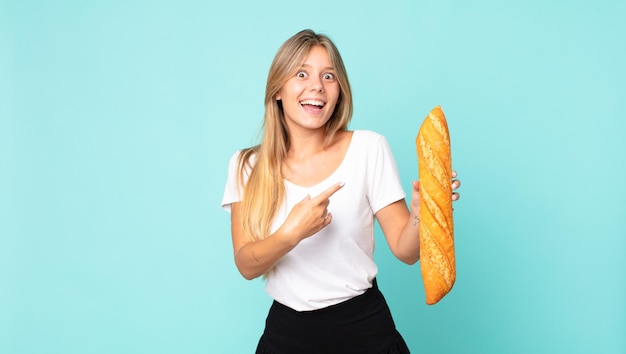 Young blonde woman looking excited and surprised pointing to the side and holding a bread baguette