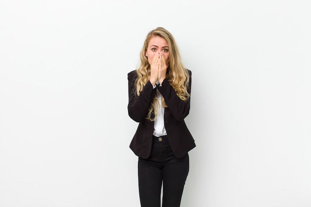 Young blonde woman looking happy, cheerful, lucky and surprised covering mouth with both hands against white wall