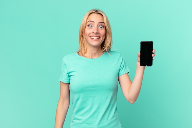 Giovane donna bionda che sembra felice e piacevolmente sorpresa e tiene in mano uno smartphone