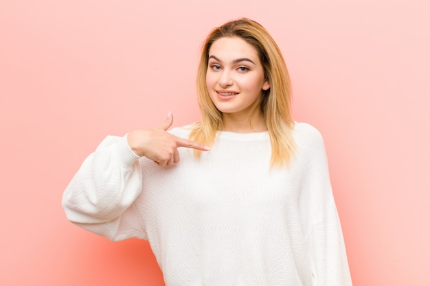 Foto giovane donna bionda che sembra felice, orgogliosa e sorpresa, che punta allegramente a se stessa, sentendosi sicura e alta sulla parete piatta rosa