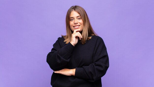 Young blonde woman looking happy and smiling with hand on chin, wondering or asking a question, comparing options