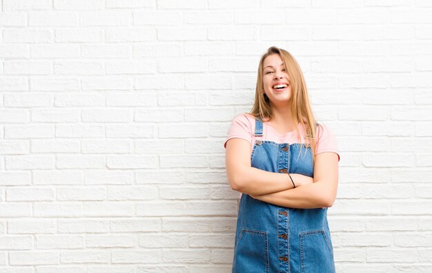 Young blonde woman looking like a happy, proud and satisfied achiever smiling with arms crossed against brick wall