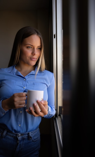 コーヒーを飲みながら窓の外を見ている若いブロンドの女性