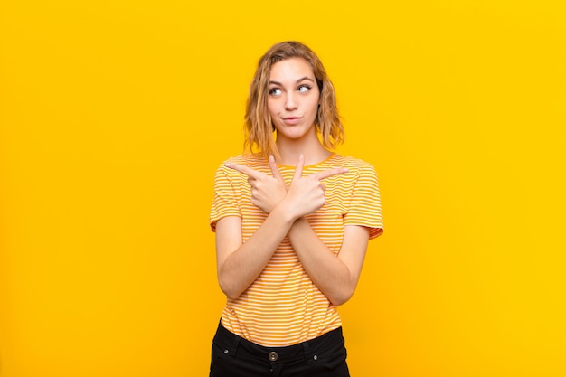 Young blonde woman looking puzzled and confused, insecure and pointing in opposite directions with doubts over wall