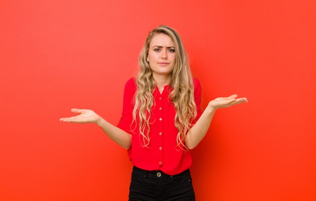 Giovane donna bionda che sembra perplessa, confusa e stressata, chiedendosi tra diverse opzioni, sentendosi incerta contro il muro rosso