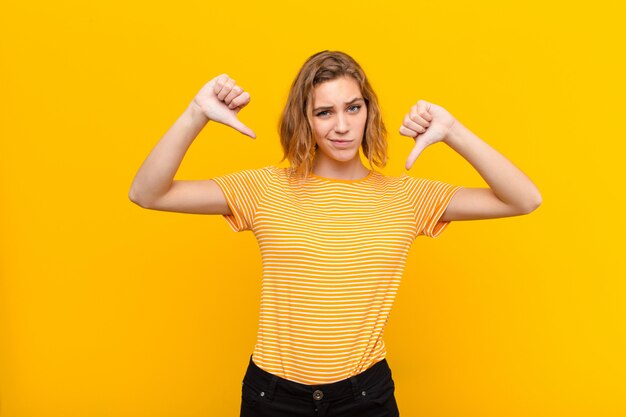 Young blonde woman looking sad, disappointed or angry, showing thumbs down in disagreement, feeling frustrated against flat color wall