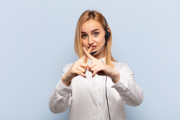 Young blonde woman looking serious and displeased with both fingers crossed up front in rejection, asking for silence