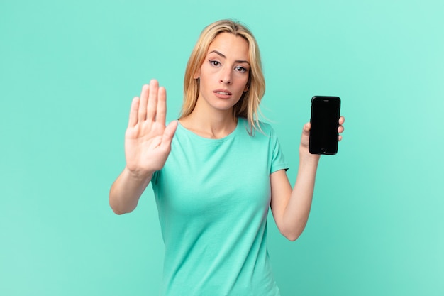 Young blonde woman looking serious showing open palm making stop gesture and holding a smart phone