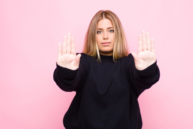 Young blonde woman looking serious, unhappy, angry and displeased forbidding entry or saying stop with both open palms against flat wall