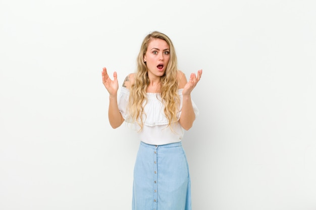 Young blonde woman looking shocked and astonished, with jaw dropped in surprise when realizing something unbelievable on white wall