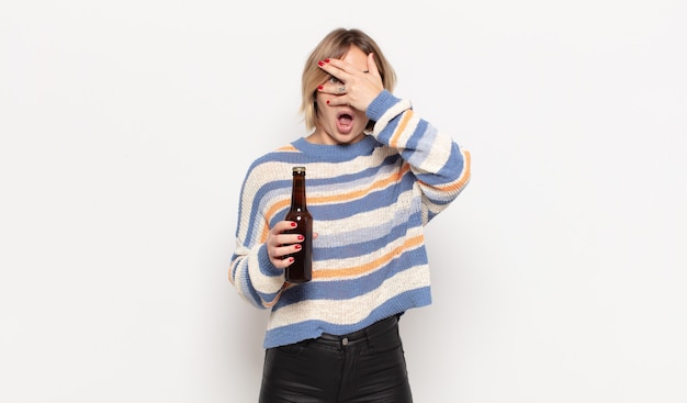 Young blonde woman looking shocked, scared or terrified, covering face with hand and peeking between fingers