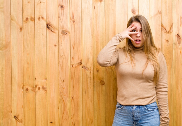 ショックを受けている、怖がっている、または恐怖を探している若いブロンドの女性、手で顔を覆い、指の間を覗く