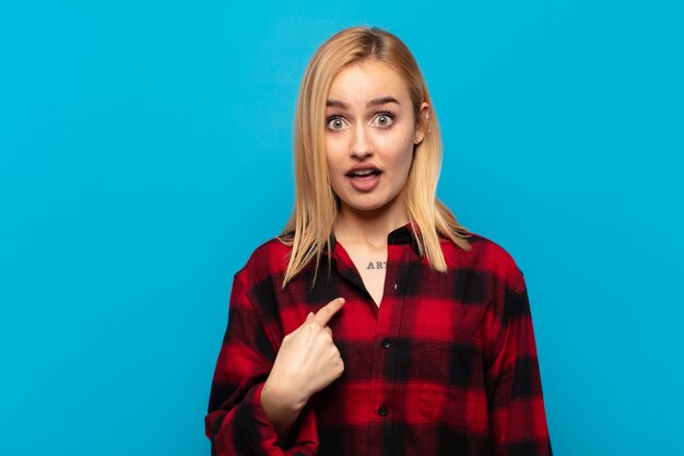 Young blonde woman looking shocked and surprised with mouth wide open, pointing to self