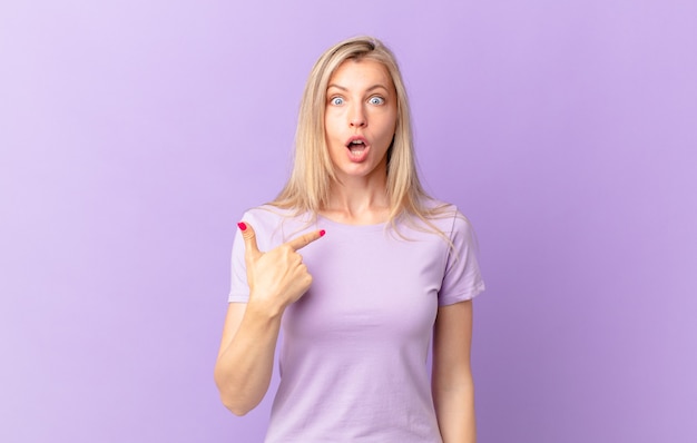 Young blonde woman looking shocked and surprised with mouth wide open, pointing to self