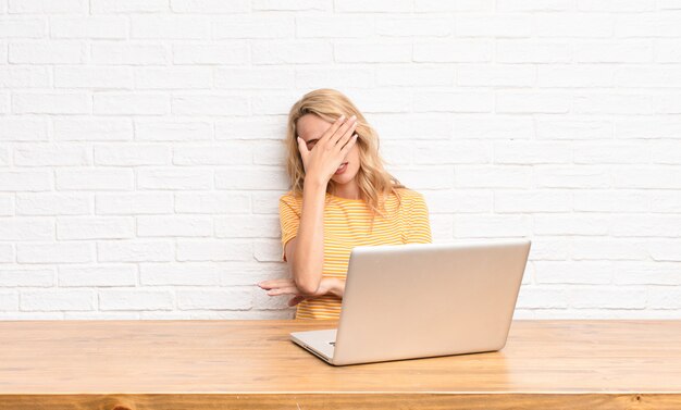 Young blonde woman looking stressed, ashamed or upset, with a headache, covering face with hand using a laptop