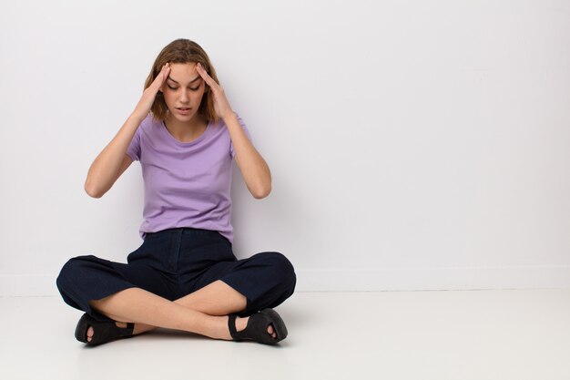 Giovane donna bionda che sembra stressata e frustrata, che lavora sotto pressione con un mal di testa e travagliata da problemi contro la parete piatta di colore