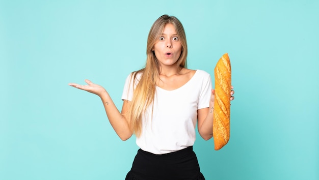 Young blonde woman looking surprised and shocked, with jaw dropped holding an object and holding a bread baguette