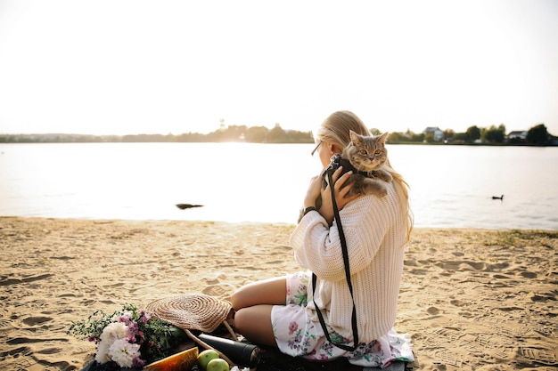 Foto giovane donna bionda sdraiata sulla spiaggia in autunno in un maglione bianco lavorato a maglia con un gatto in una giornata di sole durante un picnic