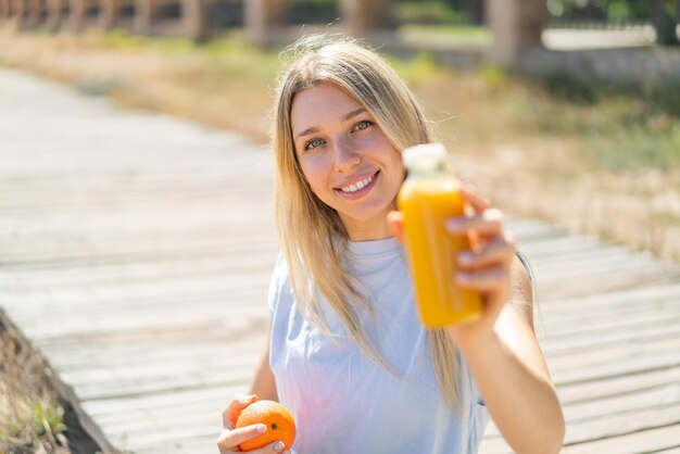 Photo young blonde woman at outdoors holding an orange and an orange juice