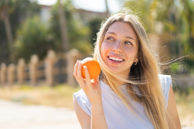 Young blonde woman at outdoors holding an orange with happy expression