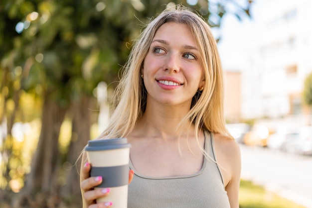 屋外で幸せな表情でテイクアウトのコーヒーを持っている若いブロンドの女性