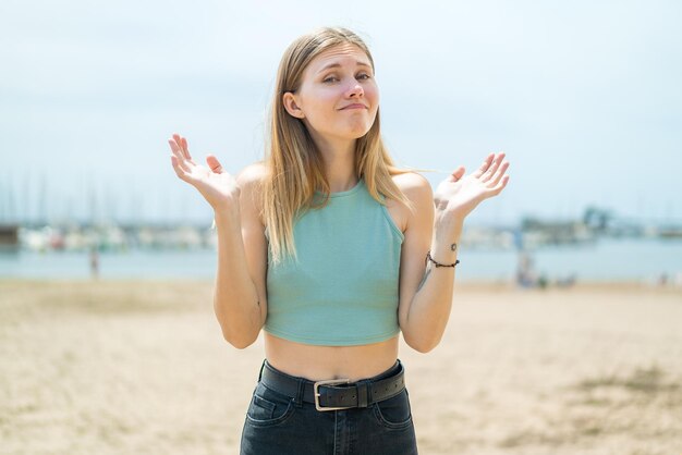 Young blonde woman at outdoors making doubts gesture