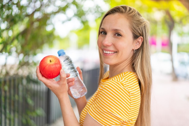 Young blonde woman at outdoors with an apple and with a bottle of water