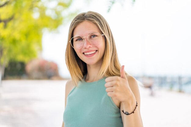 Young blonde woman at outdoors With glasses and with thumb up