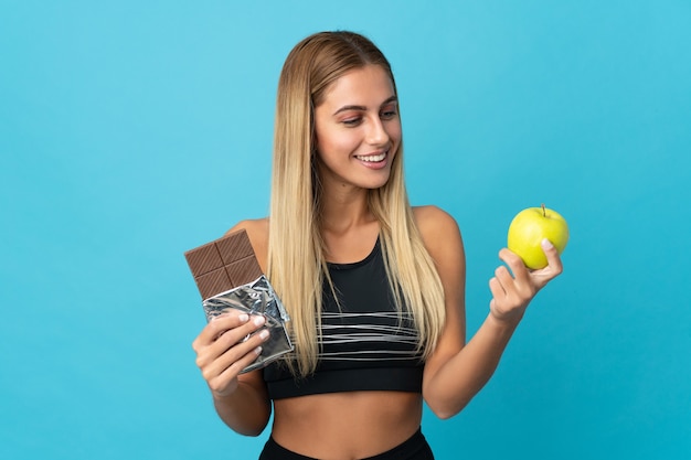 Фото Молодая блондинка в изолированном пространстве с шоколадной таблеткой в одной руке и яблоком в другой