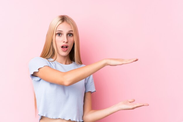 Young blonde woman on pink background shocked and amazed holding a copy space between hands.