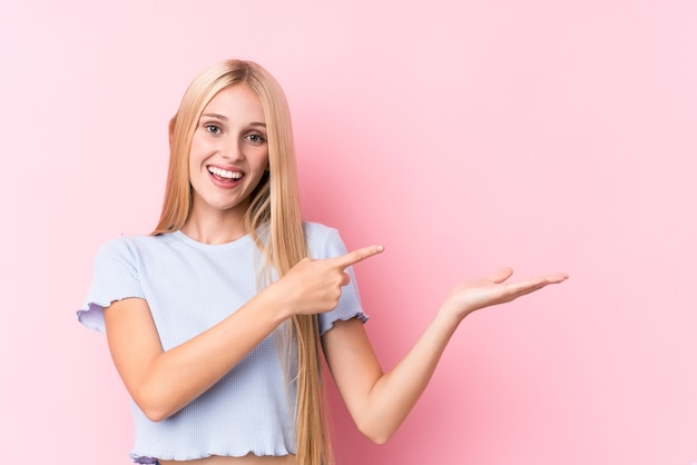 Young blonde woman on pink  excited holding a copy space on palm.