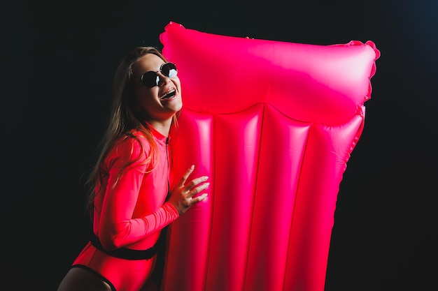 Young blonde woman in a pink swimsuit isolated on background Vacation lifestyle concept summer vacation A girl in sunglasses holds an inflatable pink mattress in her hands for swimming