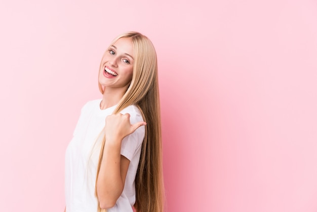 Young blonde woman on pink wall points with thumb finger away, laughing and carefree.