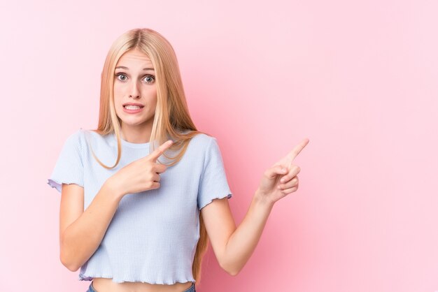 Young blonde woman on pink wall shocked pointing with index fingers to a copy space.