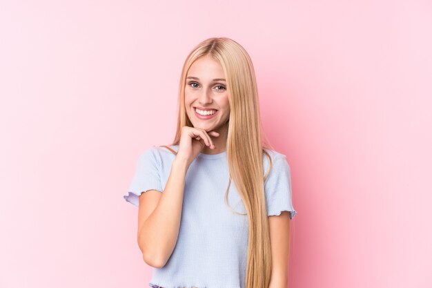 Молодая блондинка на розовой стене улыбается счастливым и уверенным, касаясь подбородка рукой.