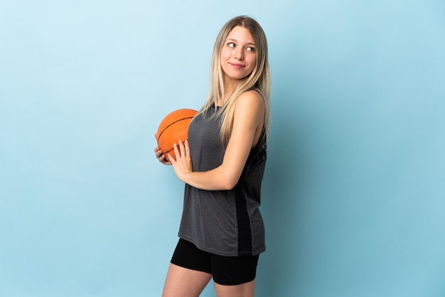 Young blonde woman playing basketball isolated on blue wall laughing