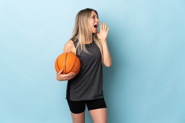 Young blonde woman playing basketball isolated on blue wall shouting with mouth wide open