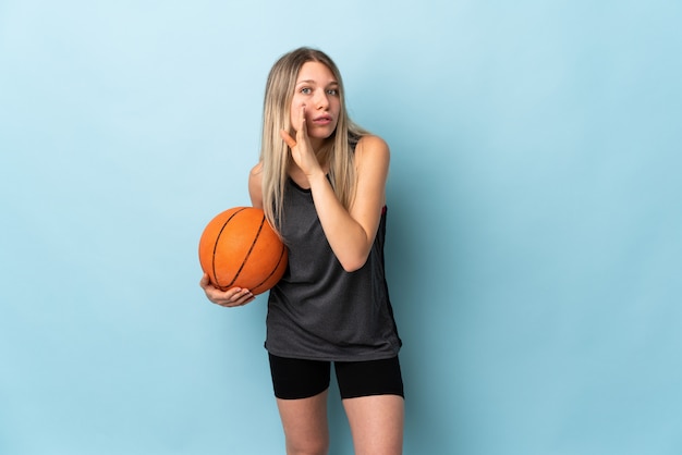 Young blonde woman playing basketball isolated on blue wall whispering something